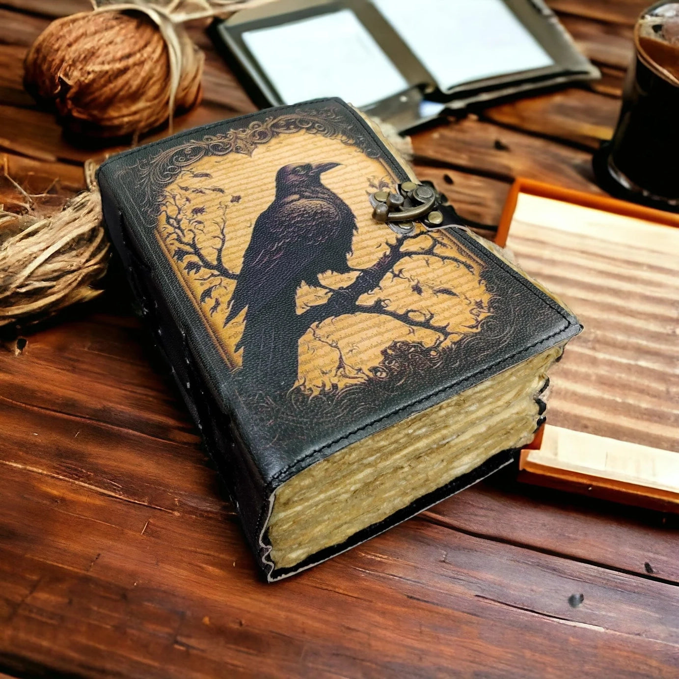 A leather journal on a wooden table. The cover depicts a drawing of a black crow perched on dark branches. Around the journal are various objects, including balls of yarn and a glass jar.