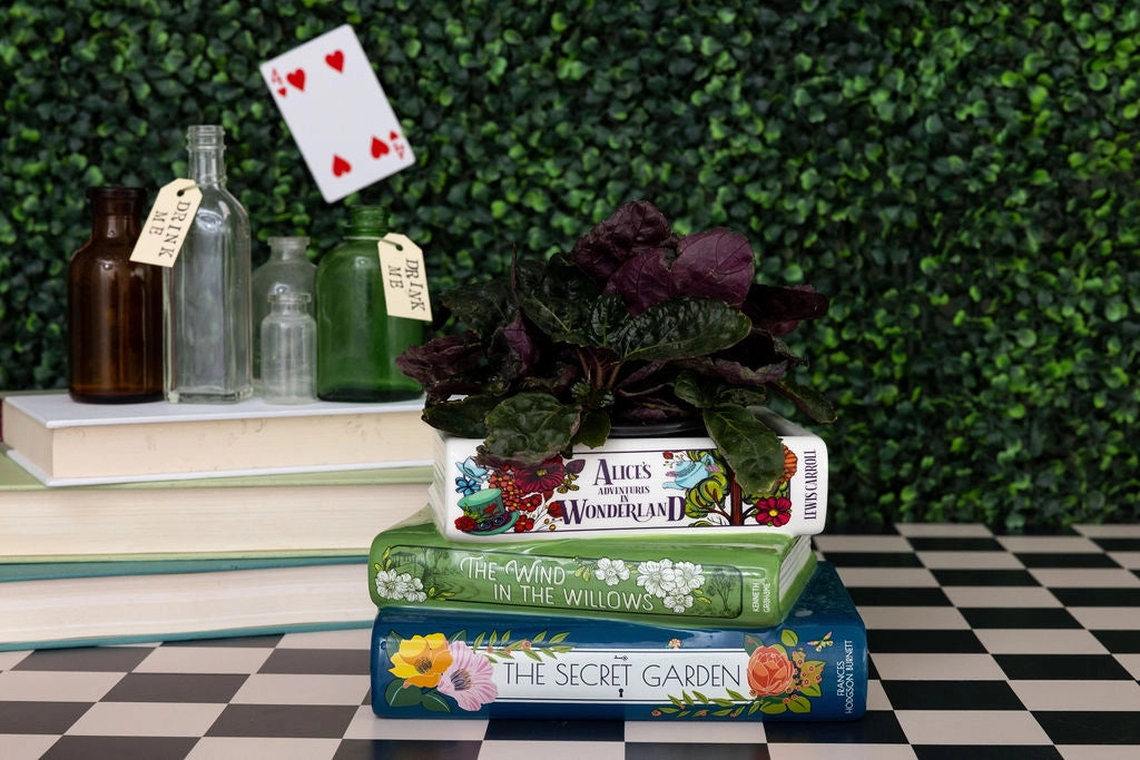 Stack of books with decorative covers on a checkered floor against a green foliage background