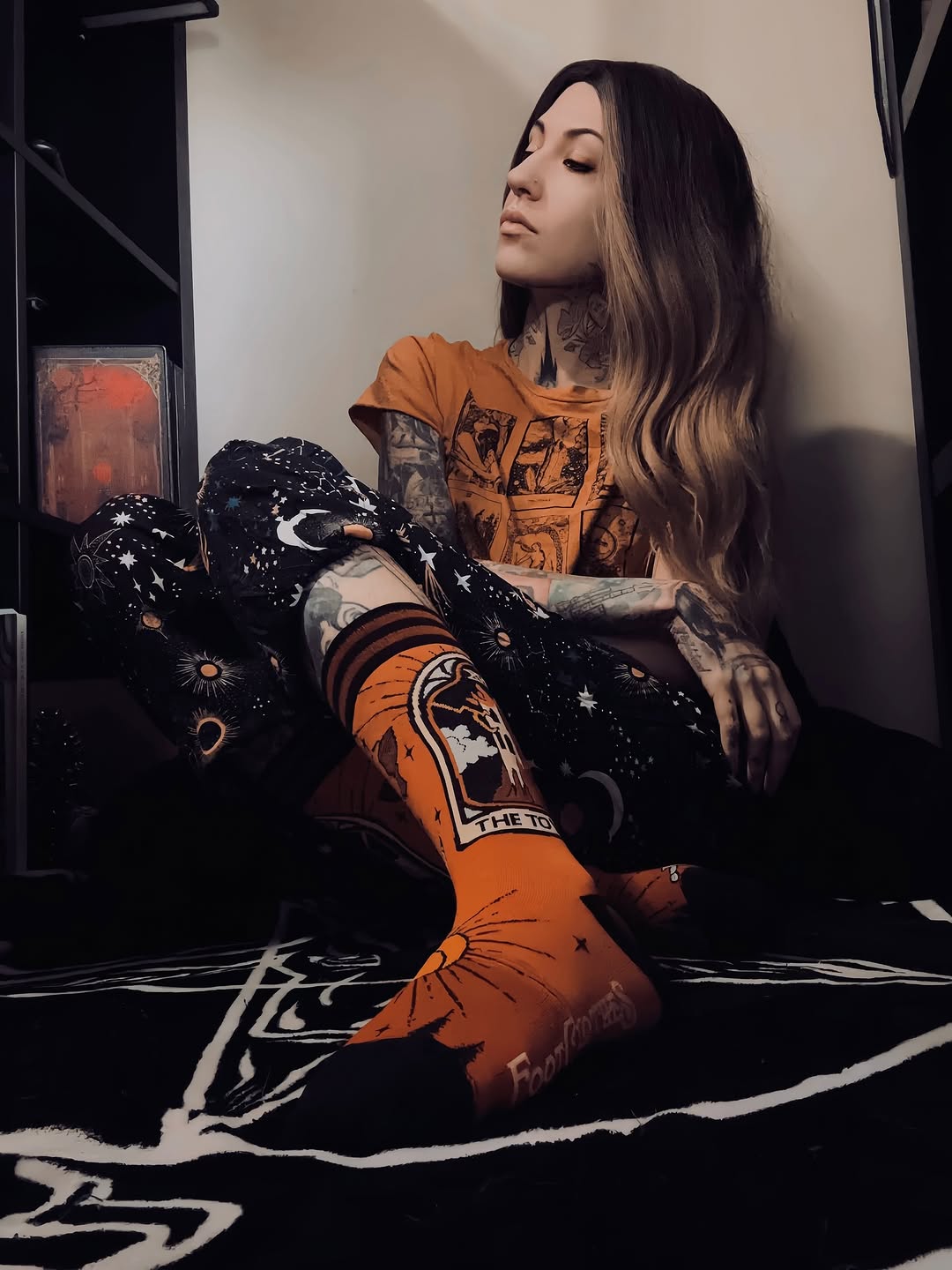 Person wearing an orange t-shirt and socks with tattoos sitting on a bed.