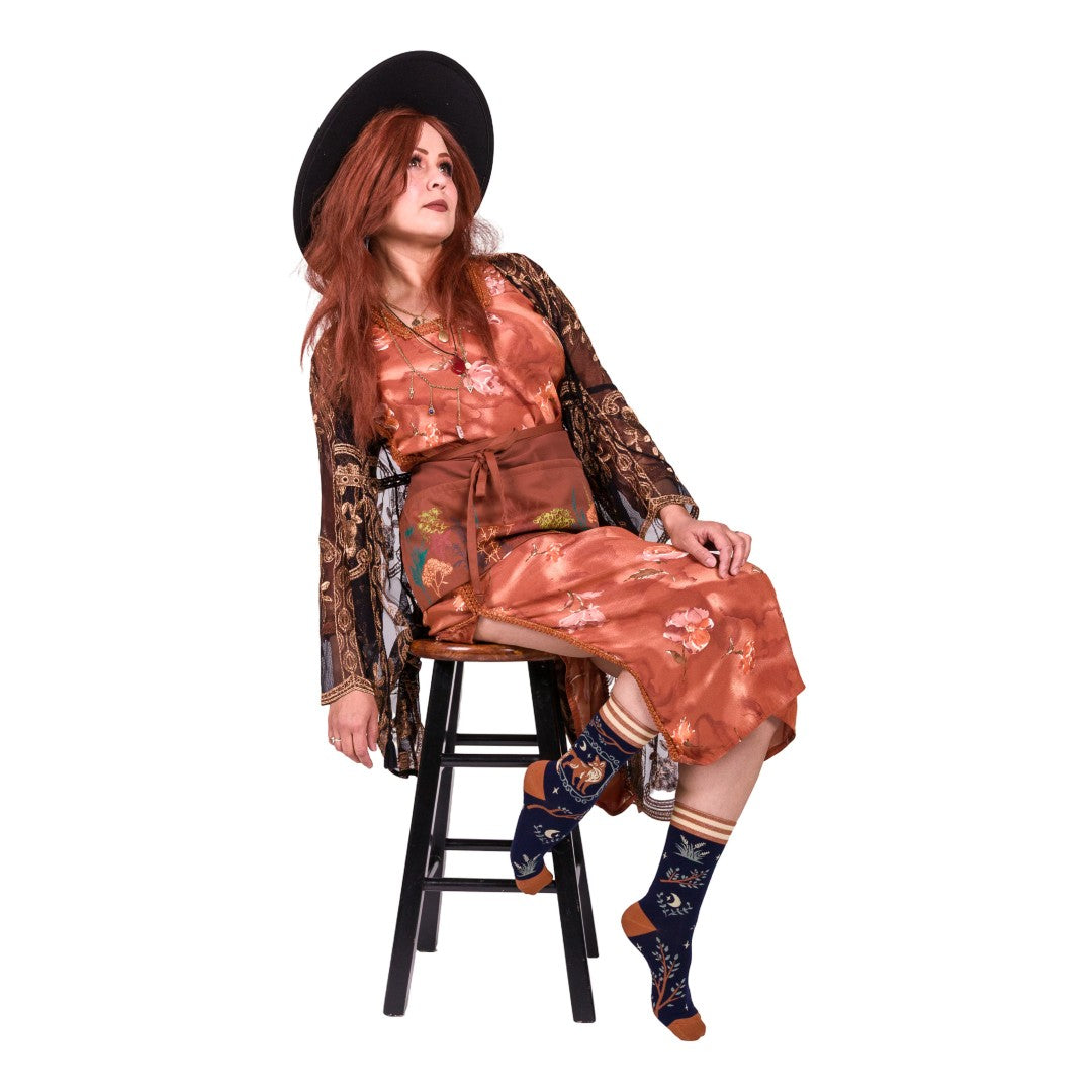 Woman sitting on a stool wearing a floral outfit with a black hat and patterned socks.