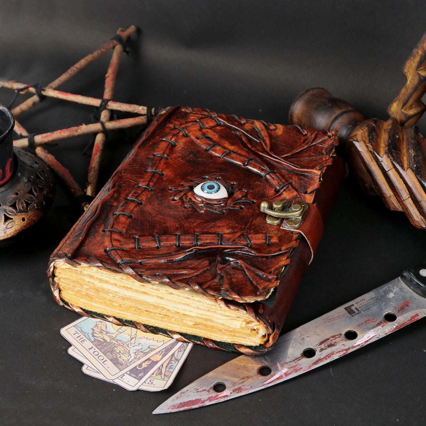 A leather journal against a grey table. The cover is styled to appear stitched together with a blue eyeball in the center. Around the journal are tarot cards, a red-stained knife and wooden trinkets.