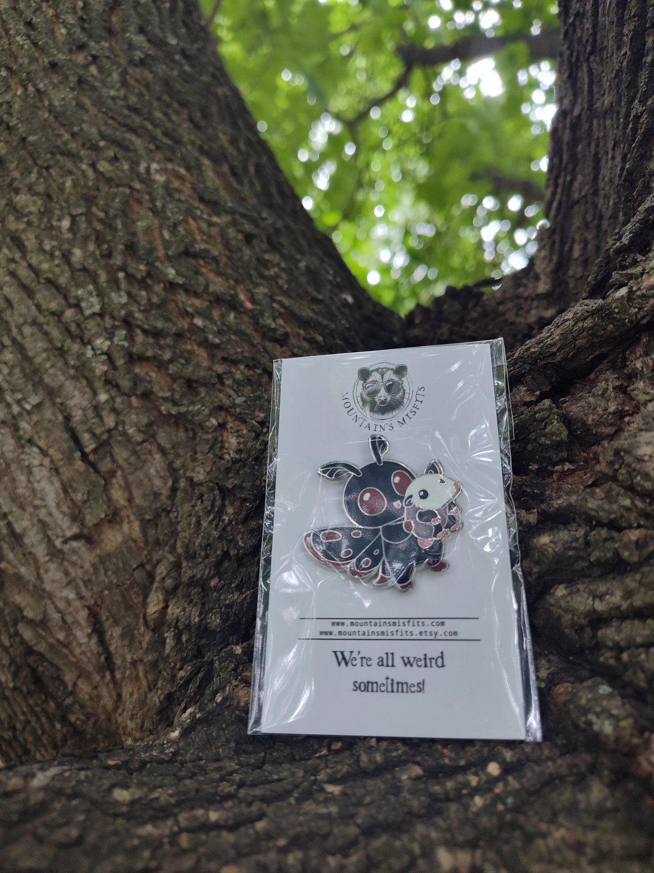 A metal pin depicting the cryptid Mothman, carrying a small oppussum. The pin is on a white backing card, with black text saying "Mountain's misfits. We're all weird sometimes." The mothman is black with red details. Behind the pin is a large tree.