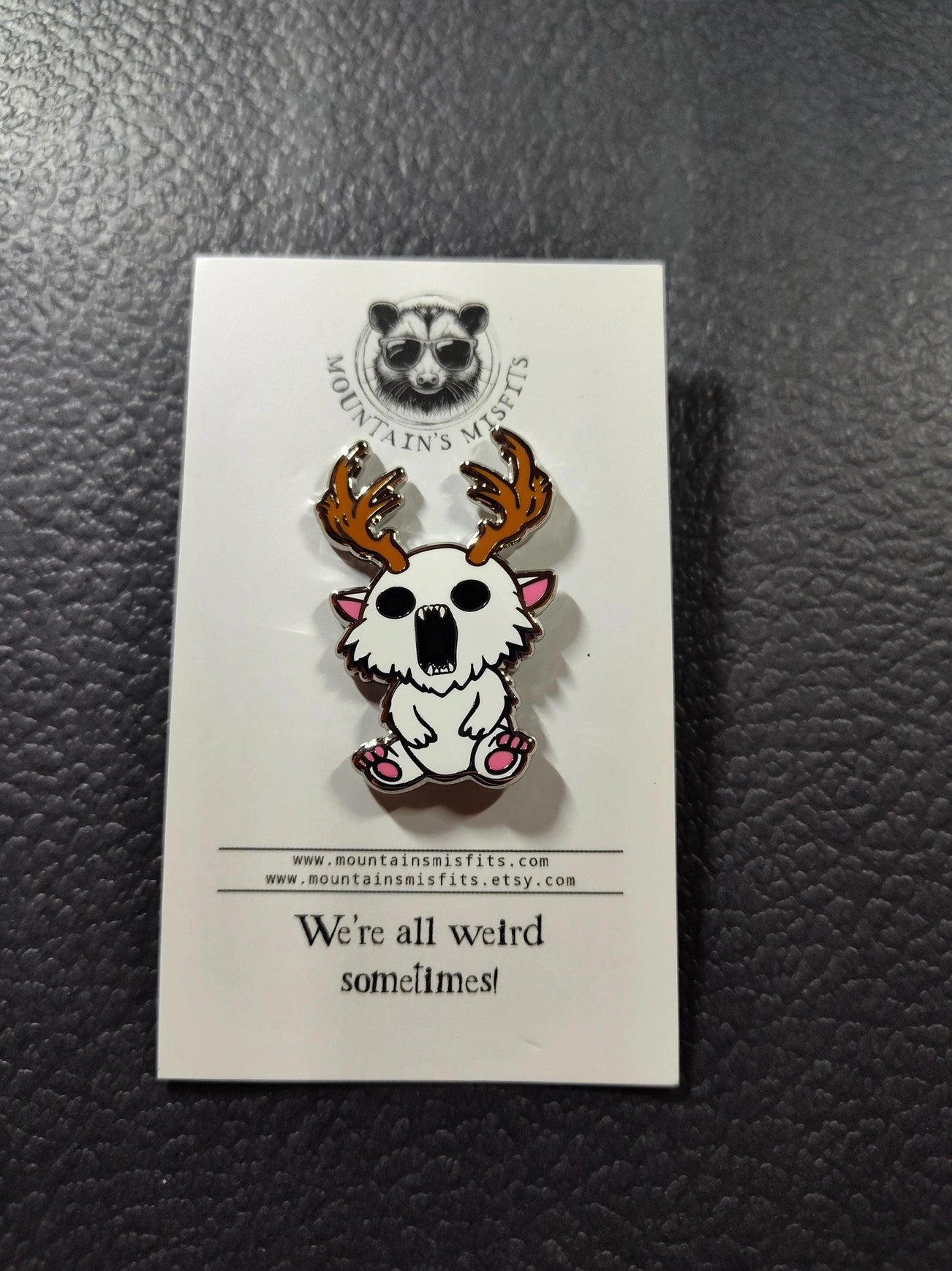 A metal pin depicting a white creature with antlers. The pin is on a white backing card, with black text saying "Mountain's misfits. We're all weird sometimes." The pin is sitting on a black leather surface.