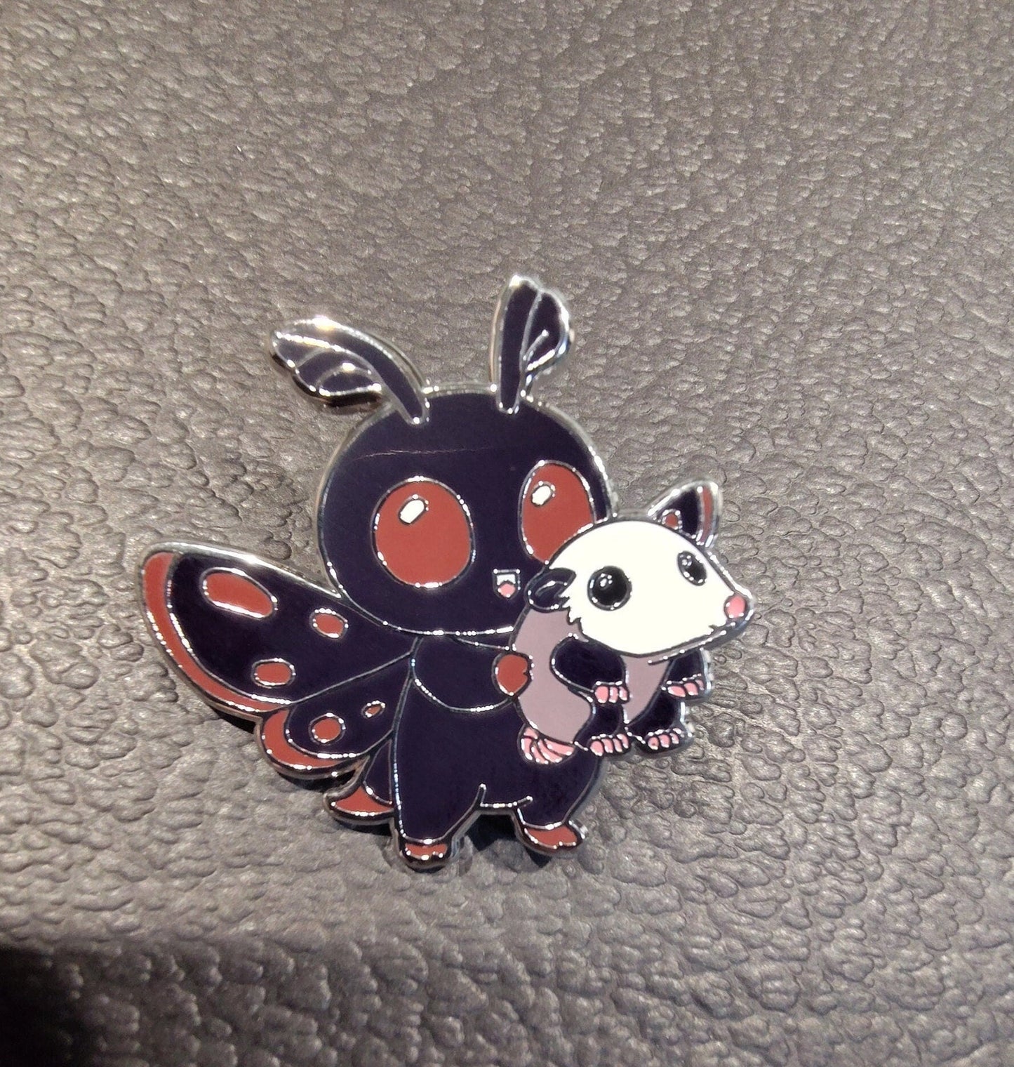 A metal pin depicting the cryptid Mothman, carrying a small oppussum. The mothman is black with red details.
