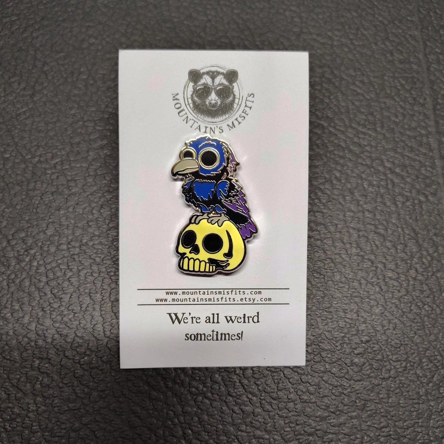 A metal pin depicting blue and grey crow, perched on a yellow skull. The pin is on a white backing card, with black text saying "Mountain's misfits. We're all weird sometimes." Behind the pin is a black leather surface.