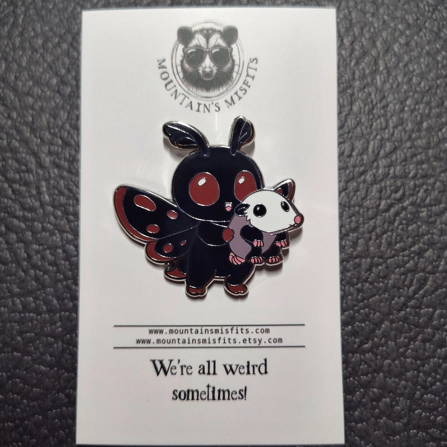 A metal pin depicting the cryptid Mothman, carrying a small oppussum. The pin is on a white backing card, with black text saying "Mountain's misfits. We're all weird sometimes." The mothman is black with red details.