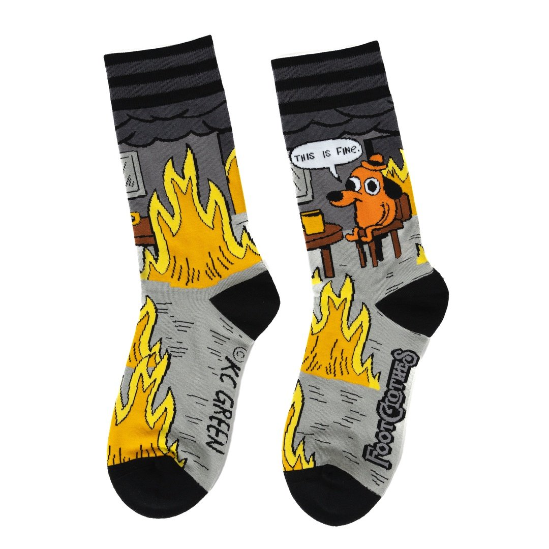 A pair of grey and back socks against a white background. One sock has a cartoon of a smilingbrown dog wearing a haat, sitting in a chair in front of burning flames. Above him is a caption saying "This is fine." The other sock has a brown wooden table with a coffee cup, and a roaring flame underneath it. The cuff and toes of the socks are black.