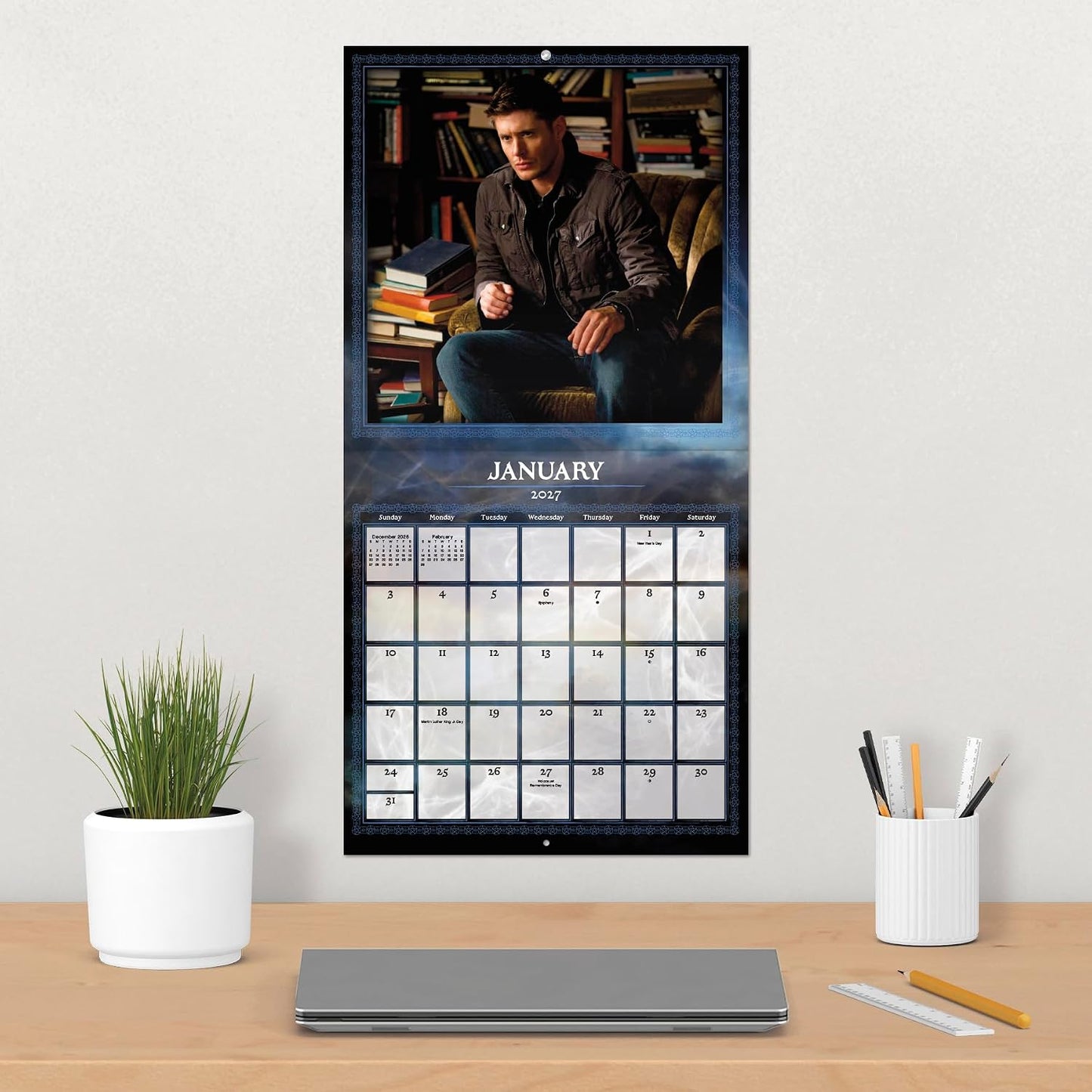 A vertical January 2024 wall calendar hanging on a light beige wall above a wooden desk. The calendar features a large portrait of a man in a dark jacket and jeans sitting in a chair in front of bookshelves, with a black-framed monthly calendar grid below.
