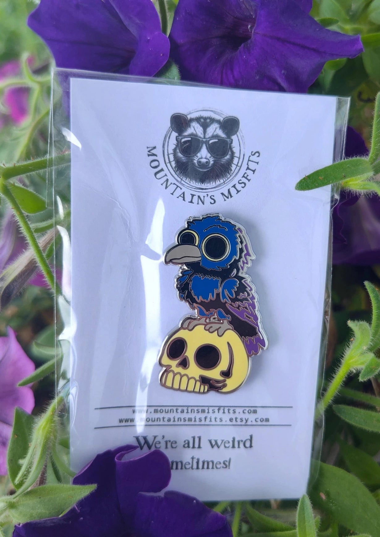 A metal pin depicting blue and grey crow, perched on a yellow skull. The pin is on a white backing card, with black text saying "Mountain's misfits. We're all weird sometimes." Behind the pin are blue and purple flowers.