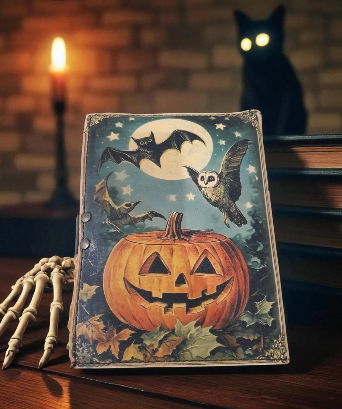A leather journal on a wooden table. The cover depicts a drawing of a large jack-o-lantern in a field, with bats and an owl flying above it against the full moon. Behind the journal is a stack of books with a black cat on top In the background is a lit candle against a brick wall.