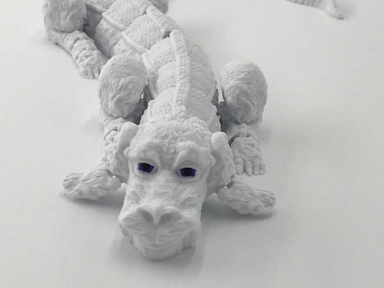 The front view of a light grey, 3D-printed rendition of Falkor the Luckdragon from The NeverEnding Story. It has dark black-purple eyes and printed joints that allow it to act as a fidget toy.