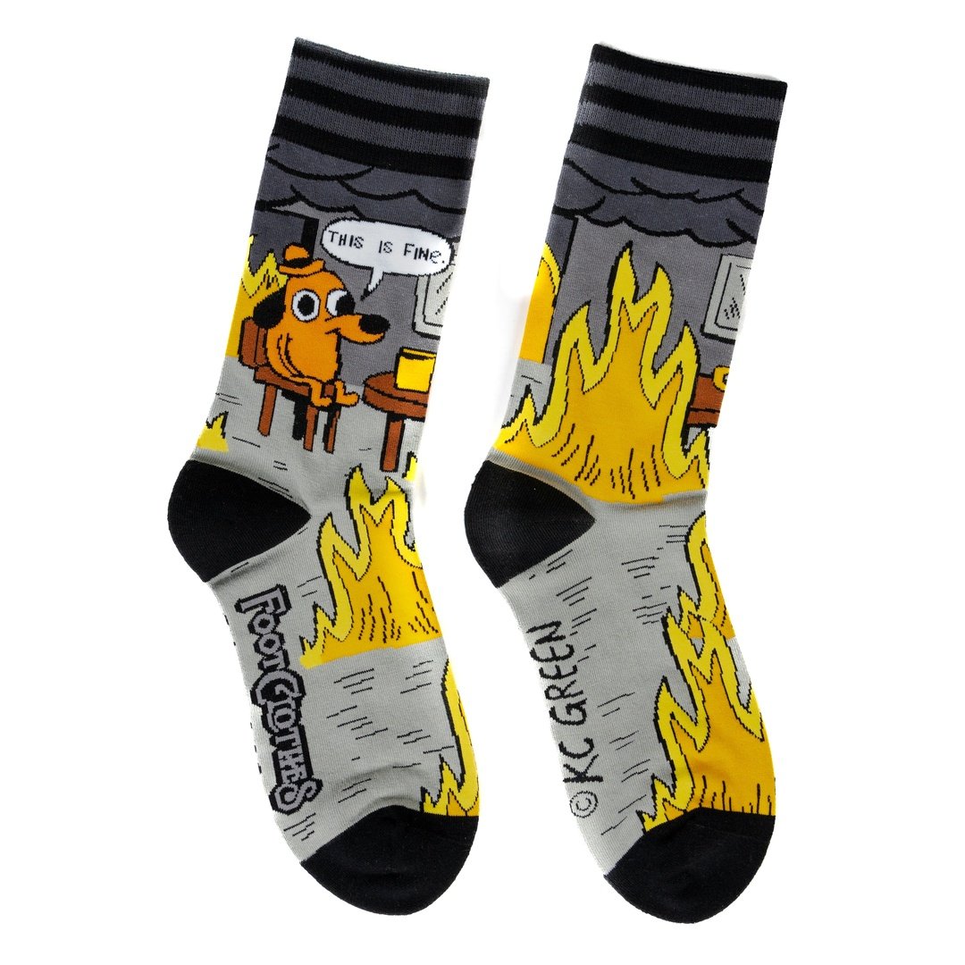 A pair of grey and back socks against a white background. One sock has a cartoon of a smilingbrown dog wearing a haat, sitting in a chair in front of burning flames. Above him is a caption saying "This is fine." The other sock has a brown wooden table with a coffee cup, and a roaring flame underneath it. The cuff and toes of the socks are black.