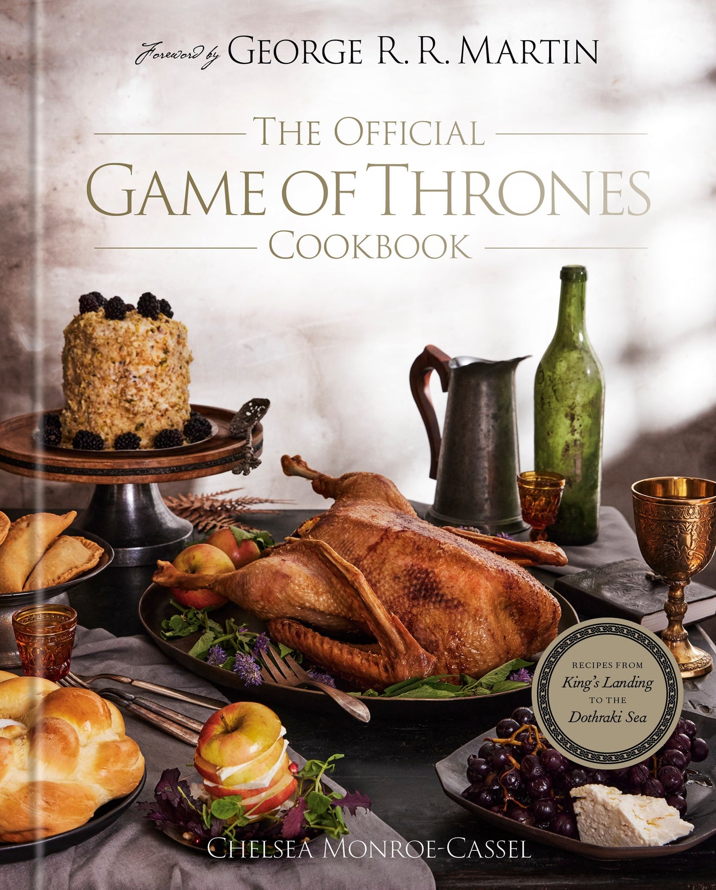 The cover of The Official Game of Thrones Cookbook. The cover features a large table with various dishes from the cookbook spread across. On the side of the table is a green wine bottle and a pewter pitcher