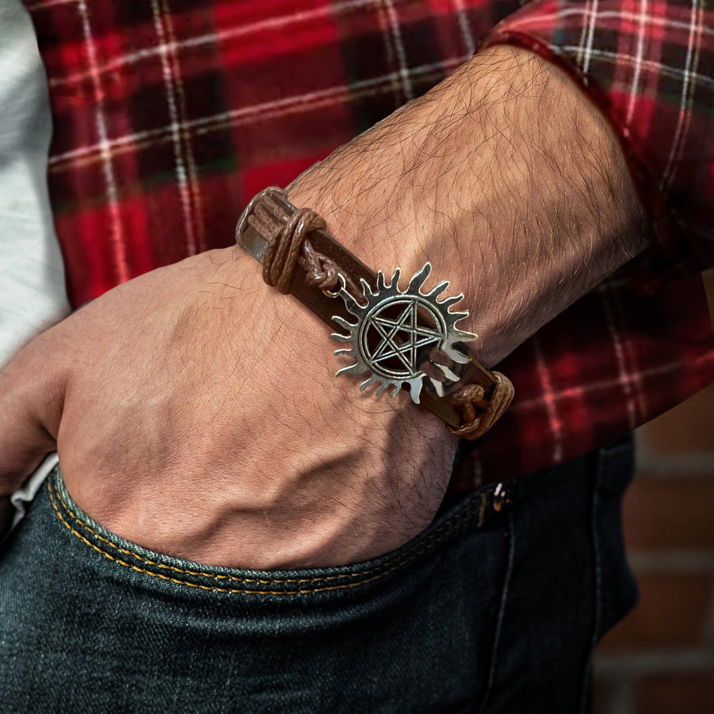 Close up of a model's wrist, wearing a brown leather bracelet with a silver charm at the top. The charm depicts the anti-possession symbol.