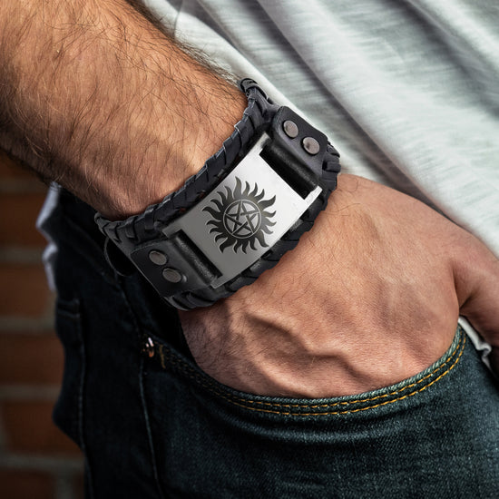 Close up view of a model's arm, with a black braided leather bracelet at the wrist. In the center of the bracelet is a metal plate with the anti-possession symbol etched onto it in black. Behind the arm is the model's white cotton shirt and blue jeans.