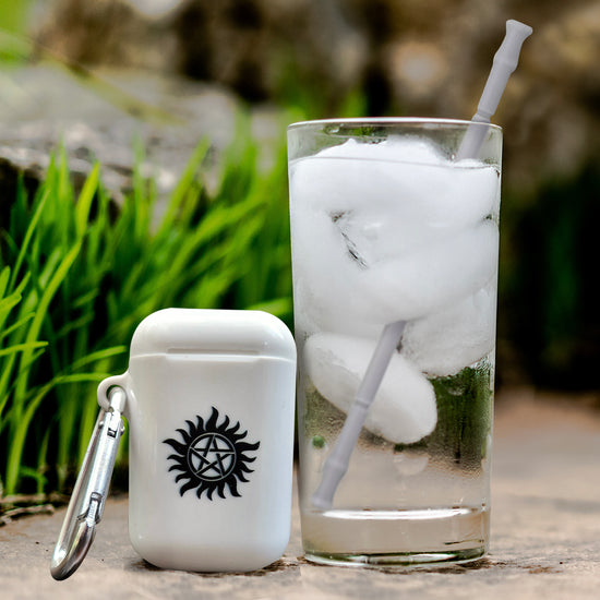 A white plastic case with the anti-possession symbol stenciled on the front in black. Next to the case is a tall glass filled with water and ice, with a grey silicone straw sticking out. Behind the case and glass are rocks and grass.