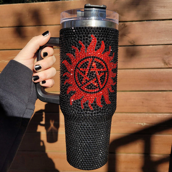 A model's hand holding a black, sparkly travel mug with a red anti-possession symbol on the front. Behind the mug is a wooden fence