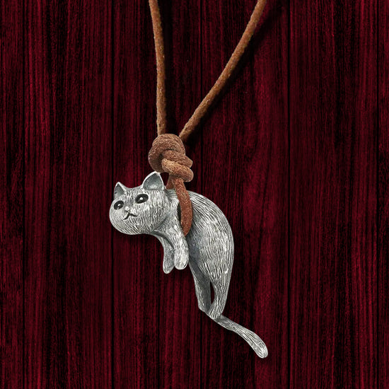 Close up of a cat-shaped pendant hanging from a brown leather cord, against a dark wooden background. The cord is wrapped under the cat's abdomen, suspending it in mid-air.