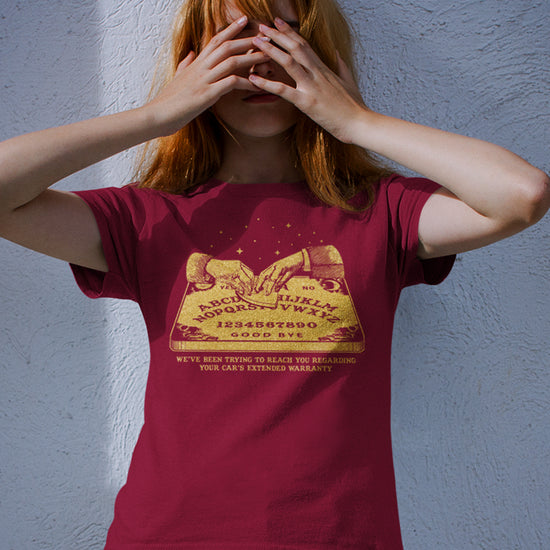 A female model wearing a maroon T-shirt, with a gold color image of two hands over a Ouija board. Under the board is gold text saying "We've been trying to reach you regarding your car's extended warranty." 