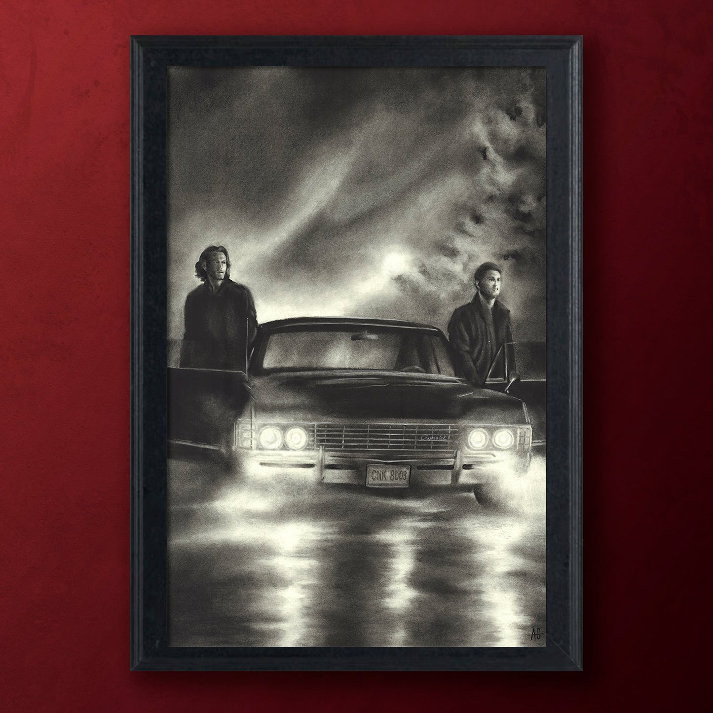A framed black and white art print depicting a 1967 Impala with two brothers standing beside it under a dramatic cloudy sky, mounted against a solid dark red wall.