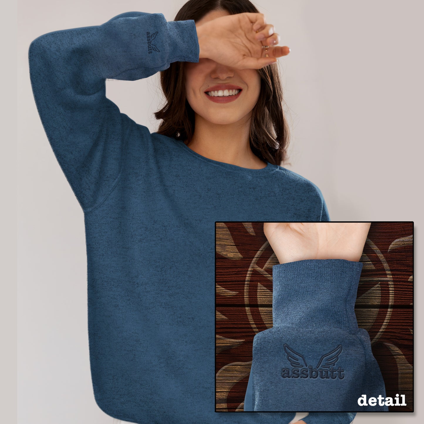 A model wearing a blue sweatshirt. An inset image shows the cuff which features an embroidered pair of wings with "assbutt" underneath