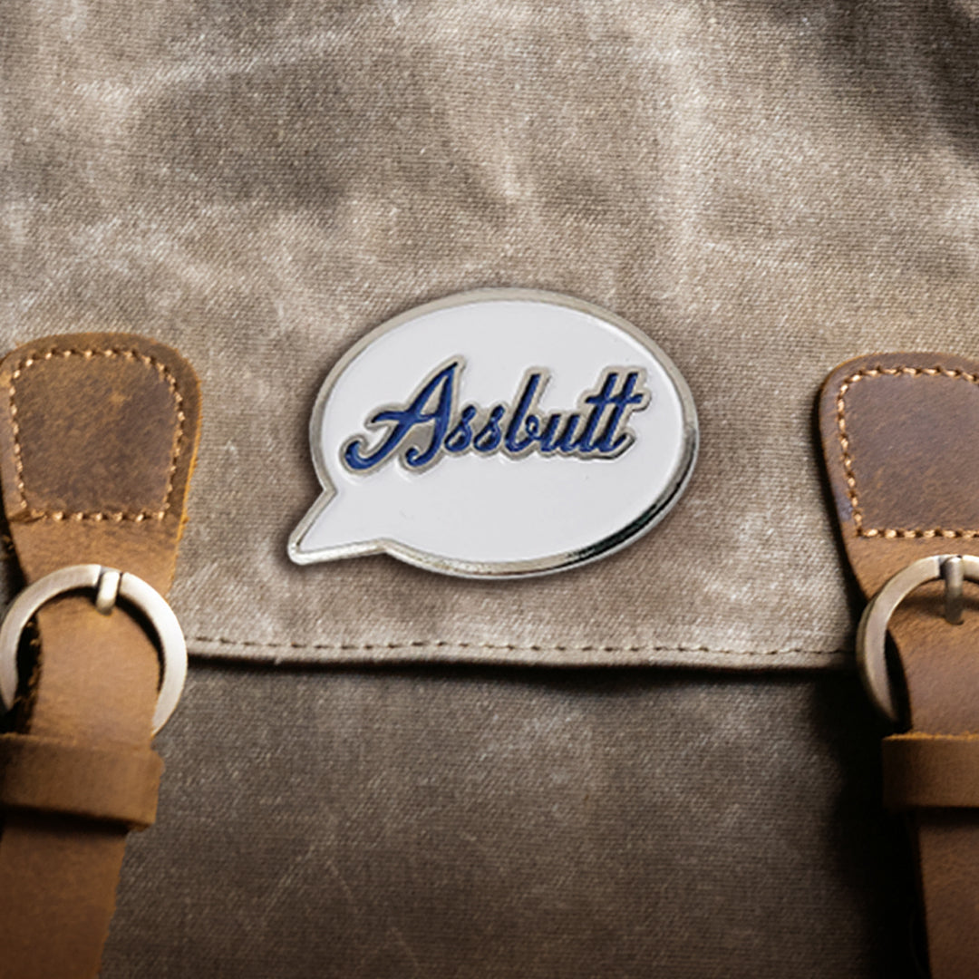 A silver pin with a white front in the shape of a speech bubble, attached to a brown canvas bag. The word "Assbutt" is written in blue script font.