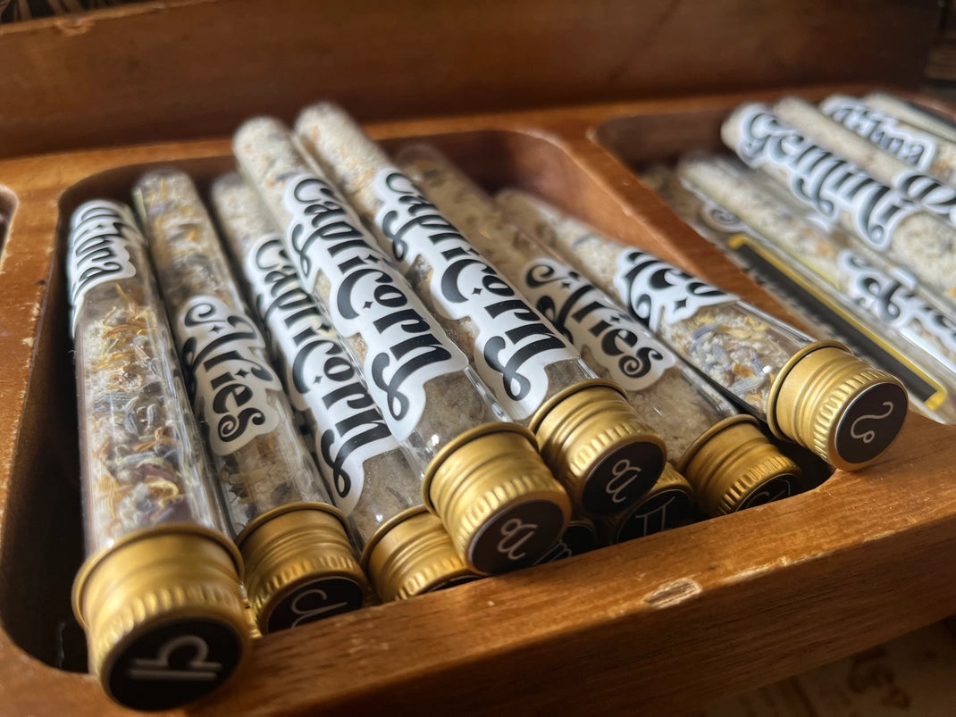 A collection of glass tubes with gold caps. Each tube has a sticker on the front, with black and white text listing the various names of the astrological signs. The tubes are sitting in a wooden box.