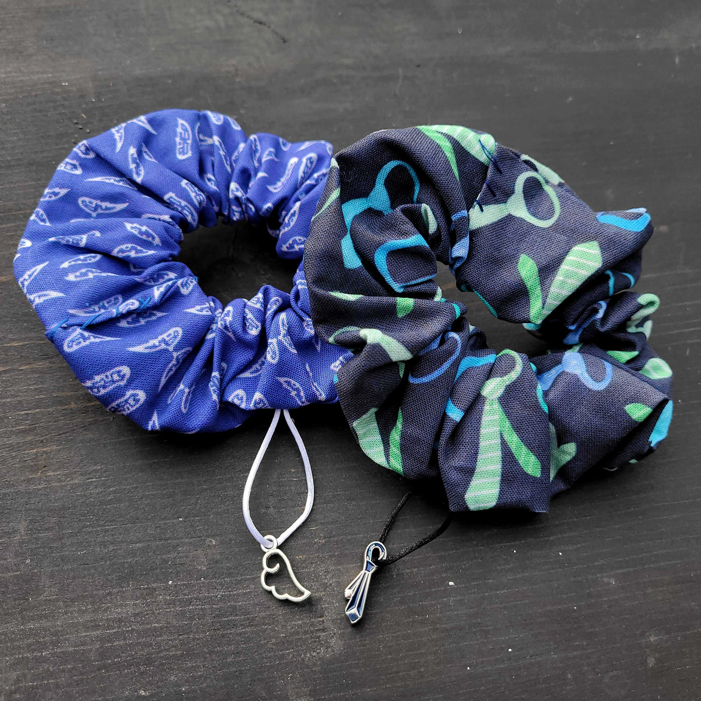 A pair of scrunchies on a black table. The left is blue with a pattern of white angel wings. The right is dark blue with a pattern of green and blue neckties.