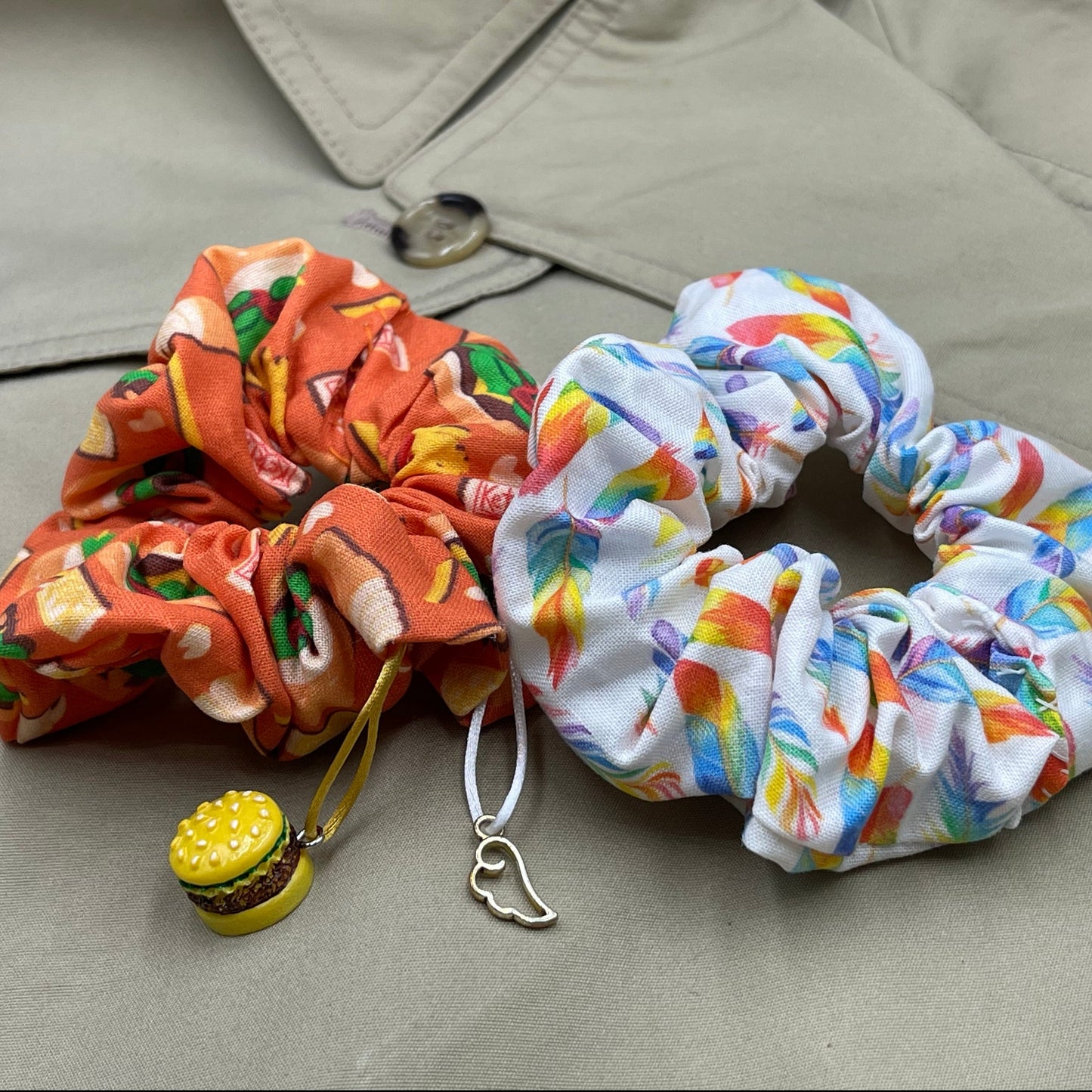 Two scrunchies side by side against a tan raincoat. The left scrunchie is orange with cartoon drawings of cheeseburgers in a pattern. A cheeseburger charm is attached to the fabric. The right scrunchie is white with rainbow colored feathers in a pattern. A silver angel wing charm is attached to the fabric