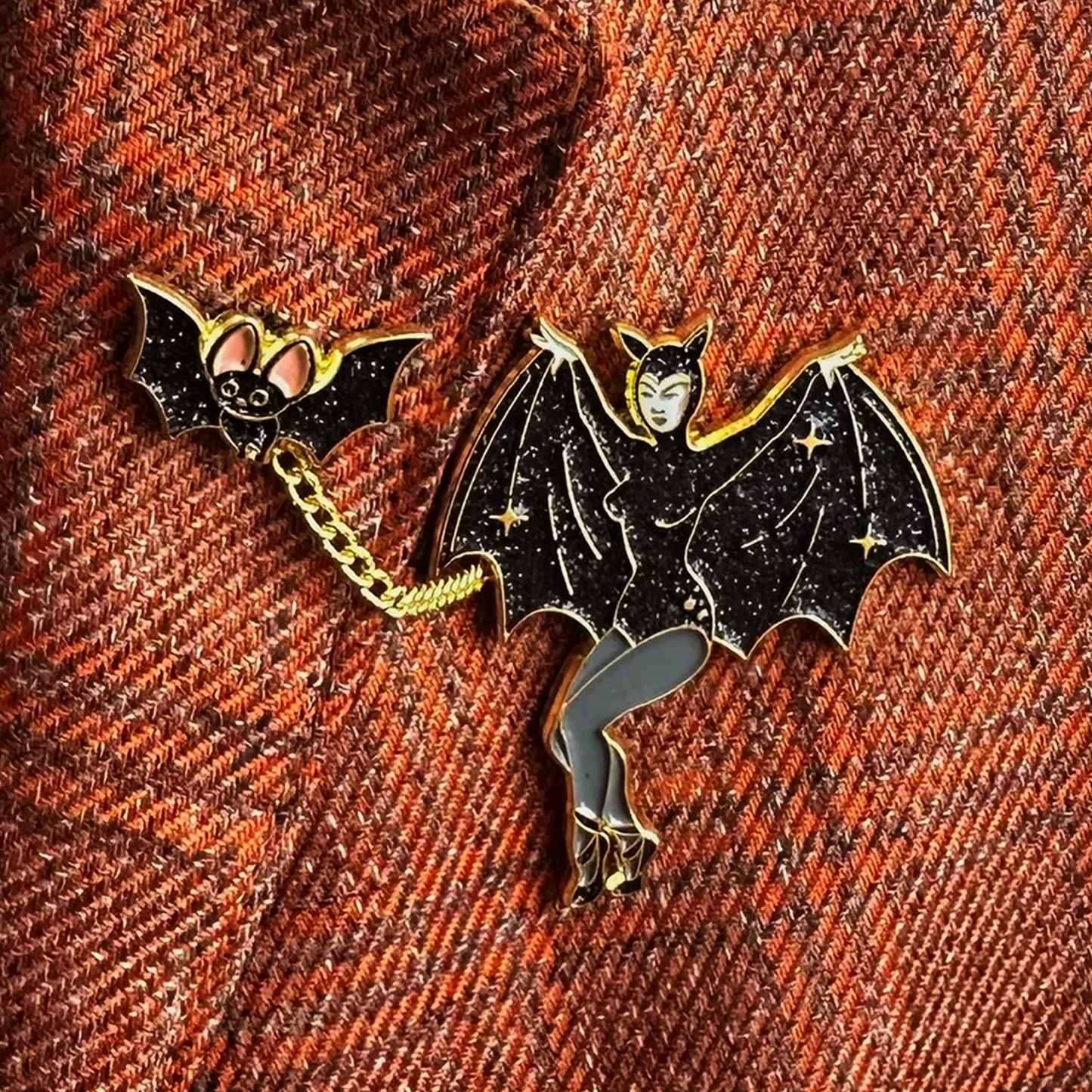 A close-up of a Halloween-themed enamel pin featuring a bat chained to a woman in a starry black costume, placed on a textured orange-brown woven fabric background.