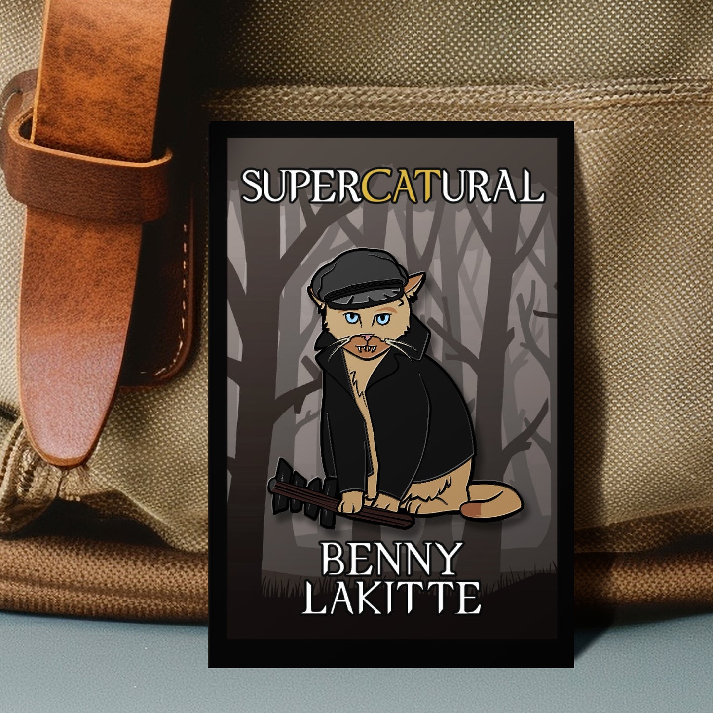 A pin a cartoon cat in a leather jacket and cap with the text 'SUPERCATURAL' and 'BENNY LAKITTE', displayed against a canvas bag with a leather strap.