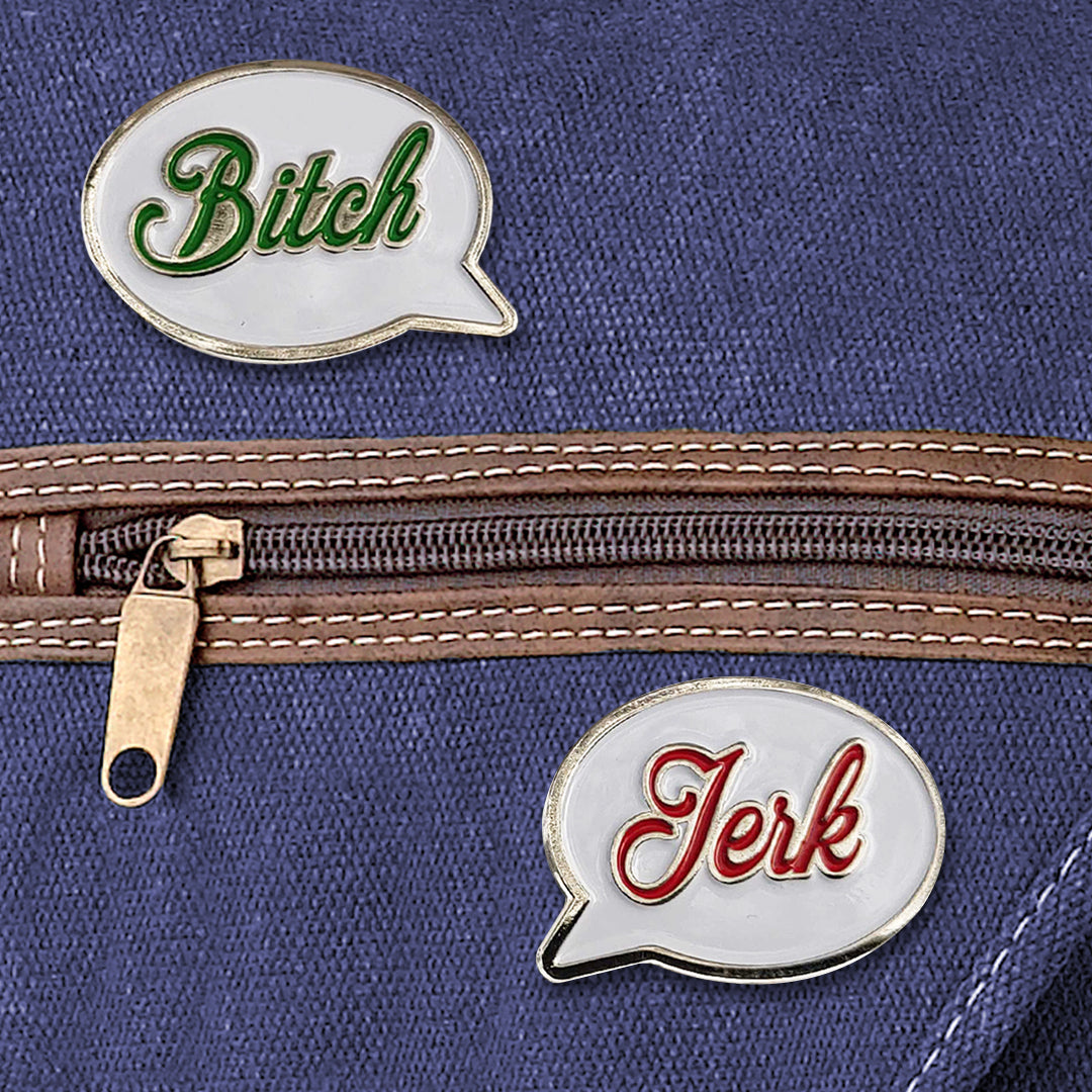 A set of two white enamel pins in the shape of speech bubbles, attached to a blue canvas bag. One reads "Bitch" in green script font. The other reads "Jerk" in red script font.