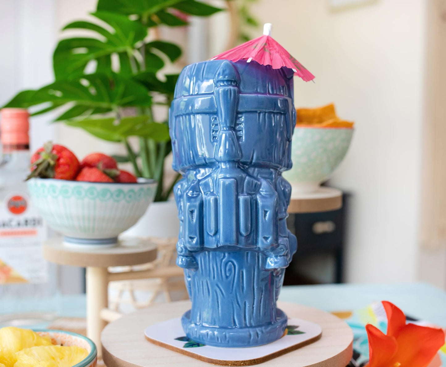 A blue tiki mug depicting the Mandalorian character Bo Katan. The mug is on a table next to bowls of fruit and a potted plant.
