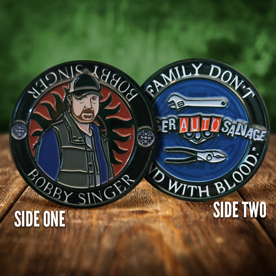 Front and back images of a challenge coin, side by side, on a wood table. The front depicts a drawing of Bobby Singer, wearing a green vest and blue shirt. Behind him is the edge of the anti-possession symbol. The back of the coin depicts a collection of tools on a blue background, and a rough sign saying "Singer auto salvage" White text around the edge says "Family don't end with blood."