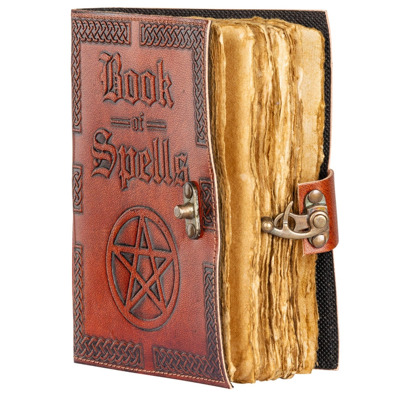 A brown leather book on a wooden table. Embossed text on the front cover says "book of spells," with an embossed pentagram symbol beneath it. On the right edge is a metal clasp to keep the book closed. Inside the book are the blank journal pages, styled to look like old parchment paper.