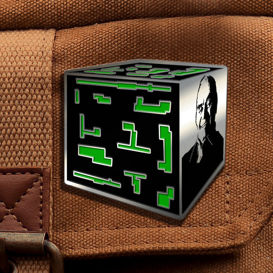 A brass pin depicting a flattened cube shape with three cube surfaces visible. Two of the surfaces are black, with random shapes in green. The last surface is a black and brass depiction of the actor Ernest Borgnine. The pin is fastened to a brown canvas bag.