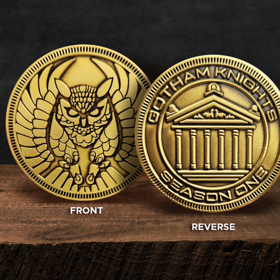 Side by side images of the front and back of a gold coin, on a wood table. The front view has an etched owl motif. The back has "Gotham Knights Season One" around the edge, with a courthouse motif etched in the center. A black background sits behind the coins.