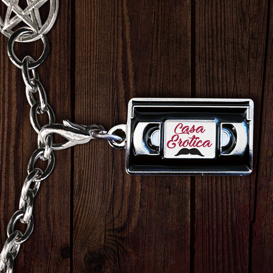 Close up of an enamel charm on a silver chain, against a dark wood table. The charm is shaped like a VHS tape, with a white label on the front. Red text says "cana erotica." A black mustache is drawn under the text.