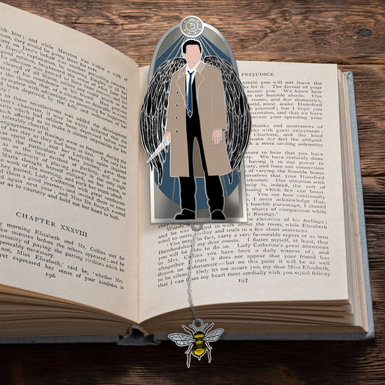 A bookmark on an open book. The bookmark depicts Castiel against a blue background. At the bottom of the bookmark is a charm attached to a chain. The charm depicts a bumblebee.