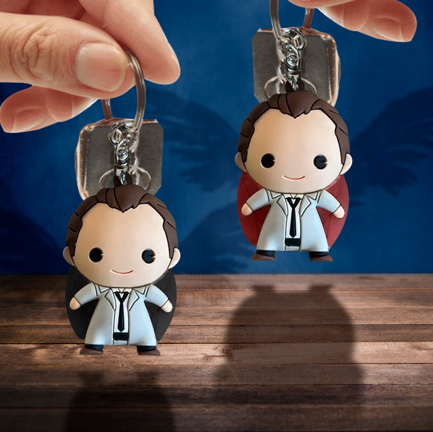 Model's hands holding up foam keychains each depicting the angel Castiel with red or black wings