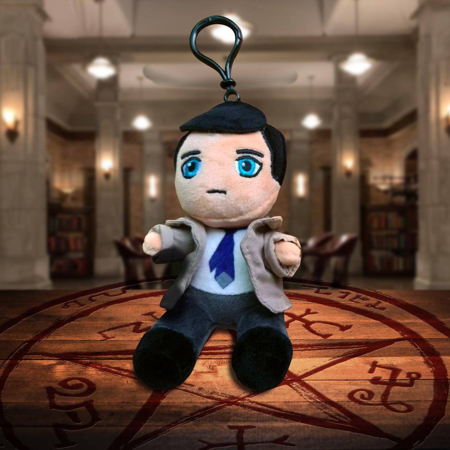A plush keychain depicting Castiel from Supernatural. Beneath the keychain is a Devil's Trap on a wooden table. In the background is The Bunker.