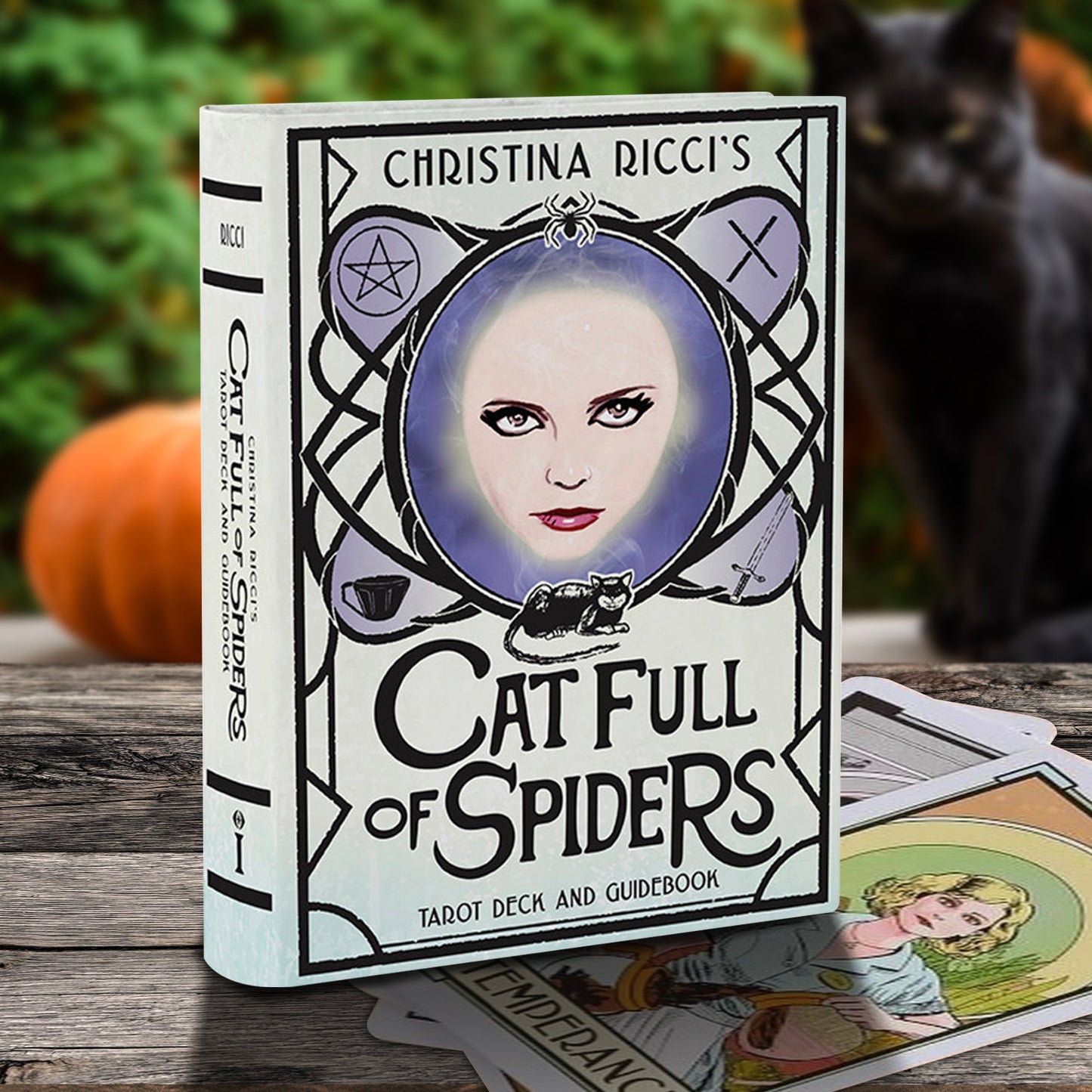 The tarot deck and guidebook 'Cat Full of Spiders' by Christina Ricci is displayed on a rustic wooden table with a pumpkin and a black cat in the background. The cover features a gothic illustration of a woman's face inside a spiderweb frame with a cat at the bottom.