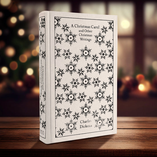 A white hardcover book on a table, in front of a lit Christmas tree. The cover has a pattern of snowflakes in black across the front. Black text at the top says "A Christmas Carol and other Christmas writings."