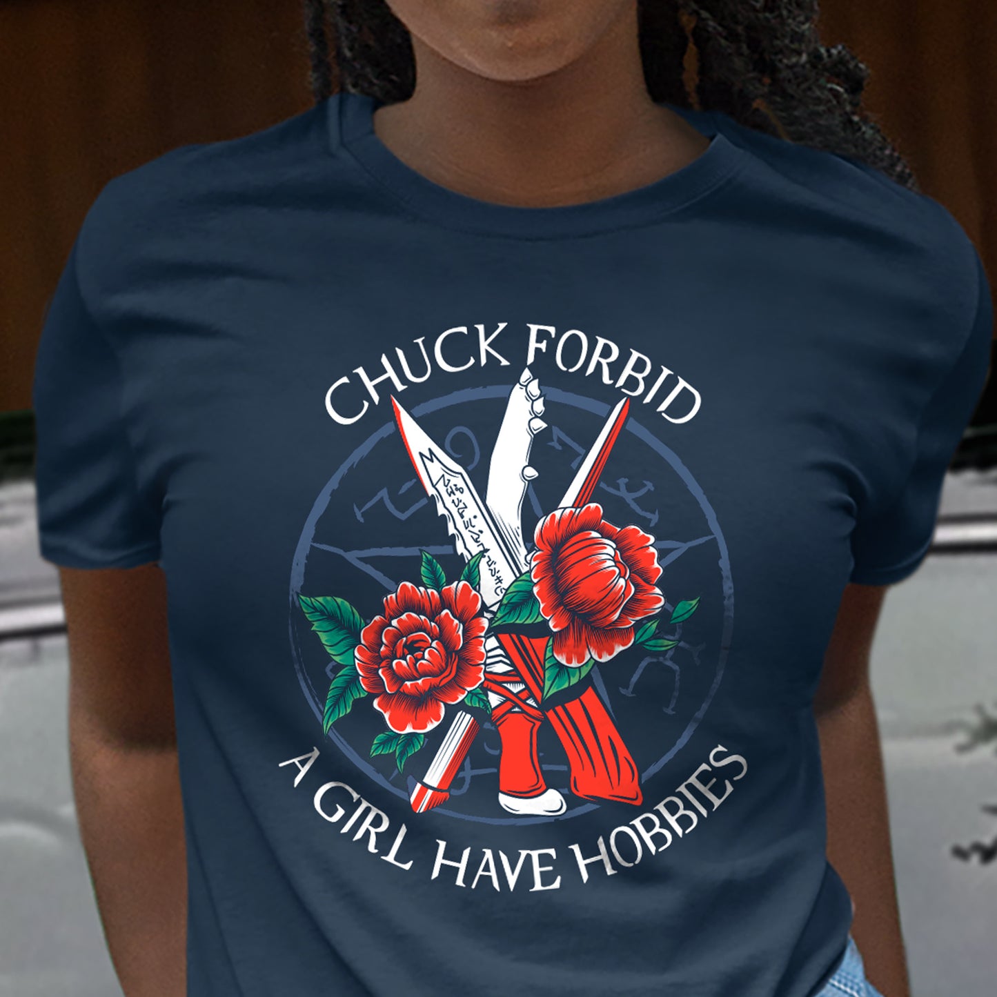A person wearing a navy graphic T-shirt. The T-shirt features a circular graphic with crossed knives, red roses, and the text 'CHUCK FORBID A GIRL HAVE HOBBIES'.