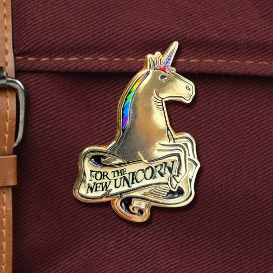 Close up of a gold colored enamel pin against a dark maroon canvas bag. The pin depicts a unicorn with a rainbow colored mane. At the center is a white scroll with black text that says "For the new unicorn."