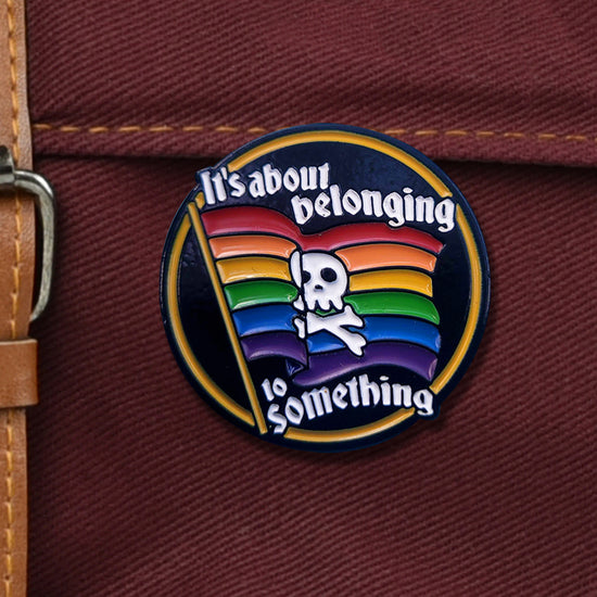 Close up view of a round enamil pin on a dark maroon canvas bag. The pin depicts a rainbow flag with a white skull and crossbones in the center. White text at the top and bottom says "it's all about belonging to something."