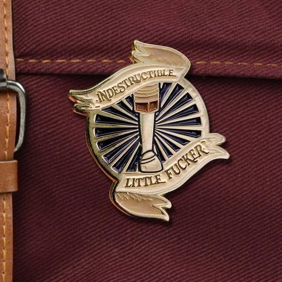 Close up of a gold colored enamel pin on a dark maroon canvas bag. The pin depicts an armored leg in front of a black background with sunbeans radiating from the center. Ribbon shaped banners at the top and bottom have black text saying "indestructible little fucker."