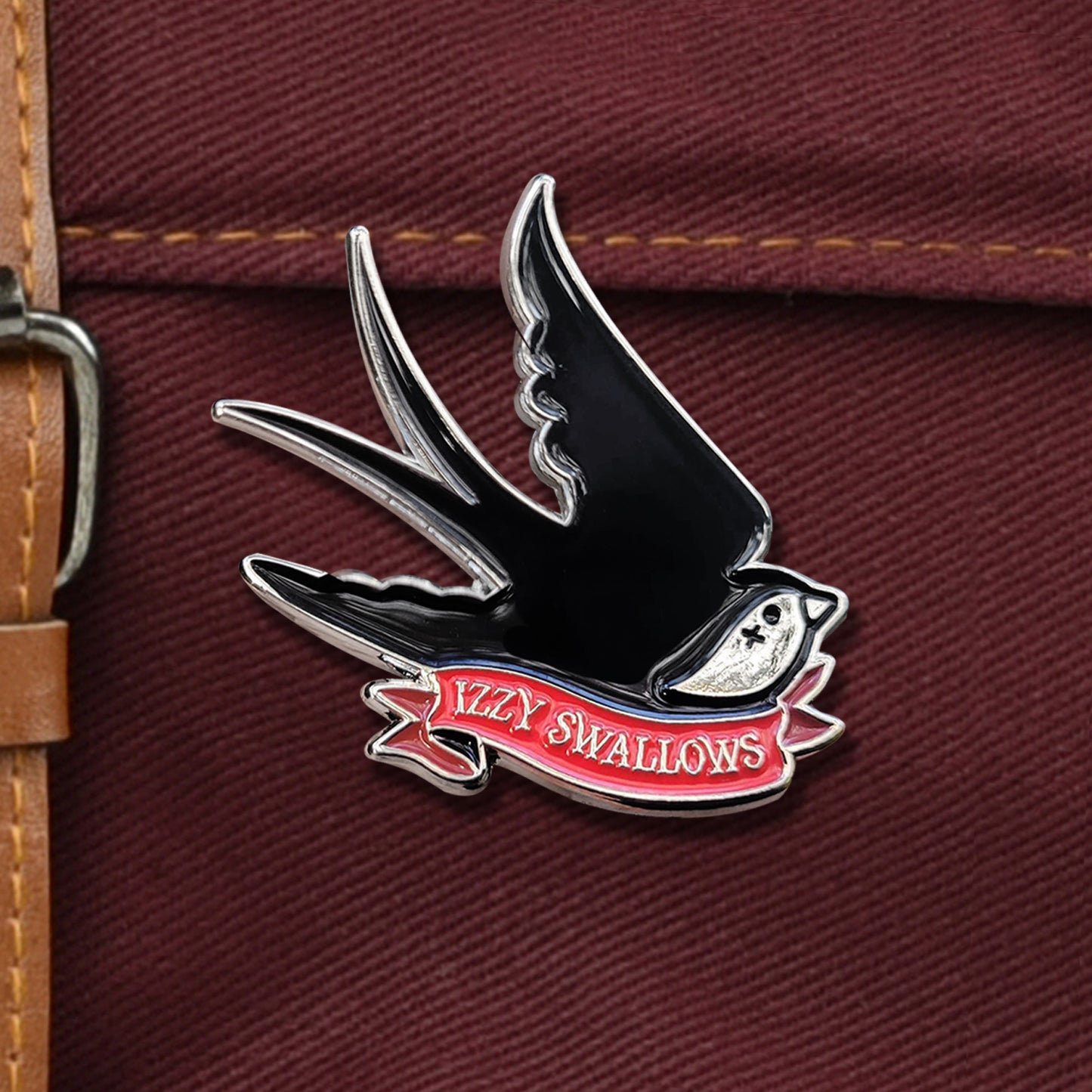 Close up of a black enamel pin shaped like a bird with a white face, against a dark maroon canvas bag. At the bird's bottom is a red ribbon with silver text that says "izzy swallows."