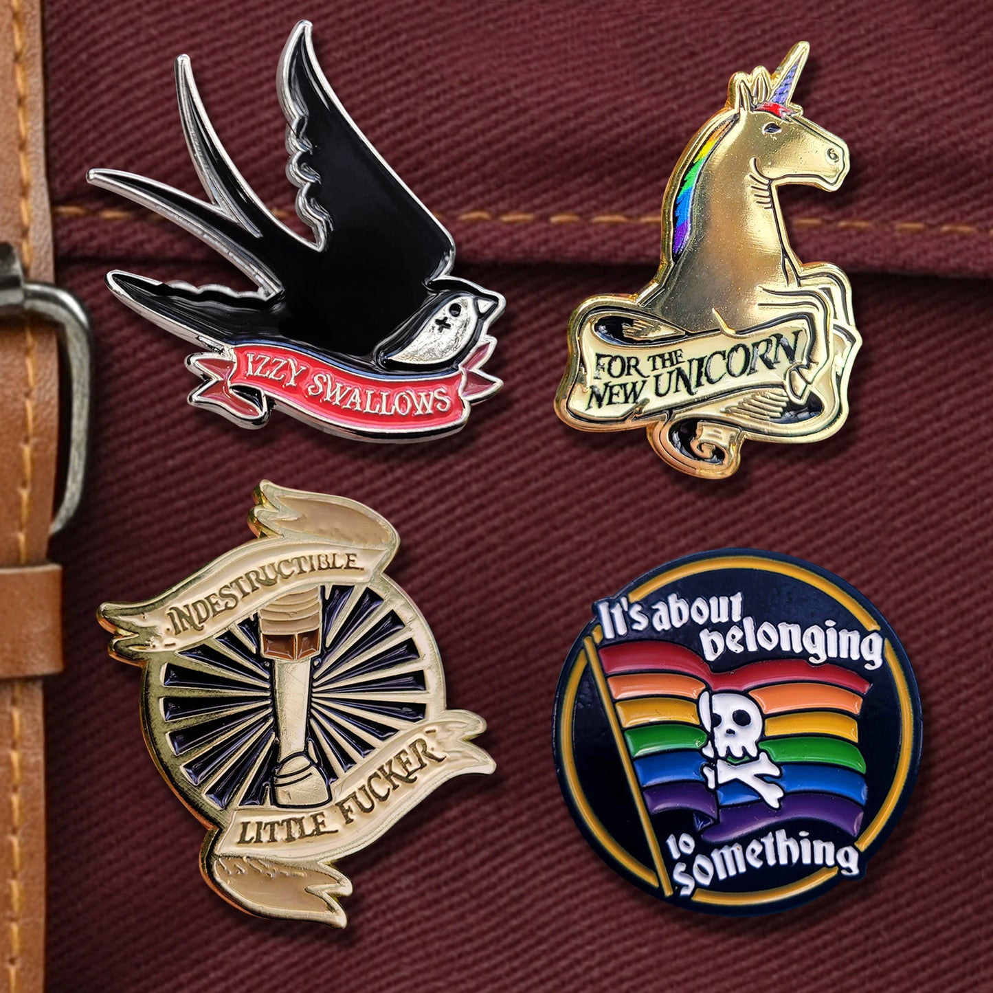 Four enamel pins against a maroon canvas bag. Top left pin is shaped like a black bird, with a red banner saying "Izzy Swallows." Top right is a unicorn with a rainbow mane, and a banner saying "for the new unicorn." Botton left is a wooden leg surrounded by sun rays, and a banner saying "indestructible little fucker" and bottom right is a rainbow skull and crossbones flaag saying it's about belonging to something.