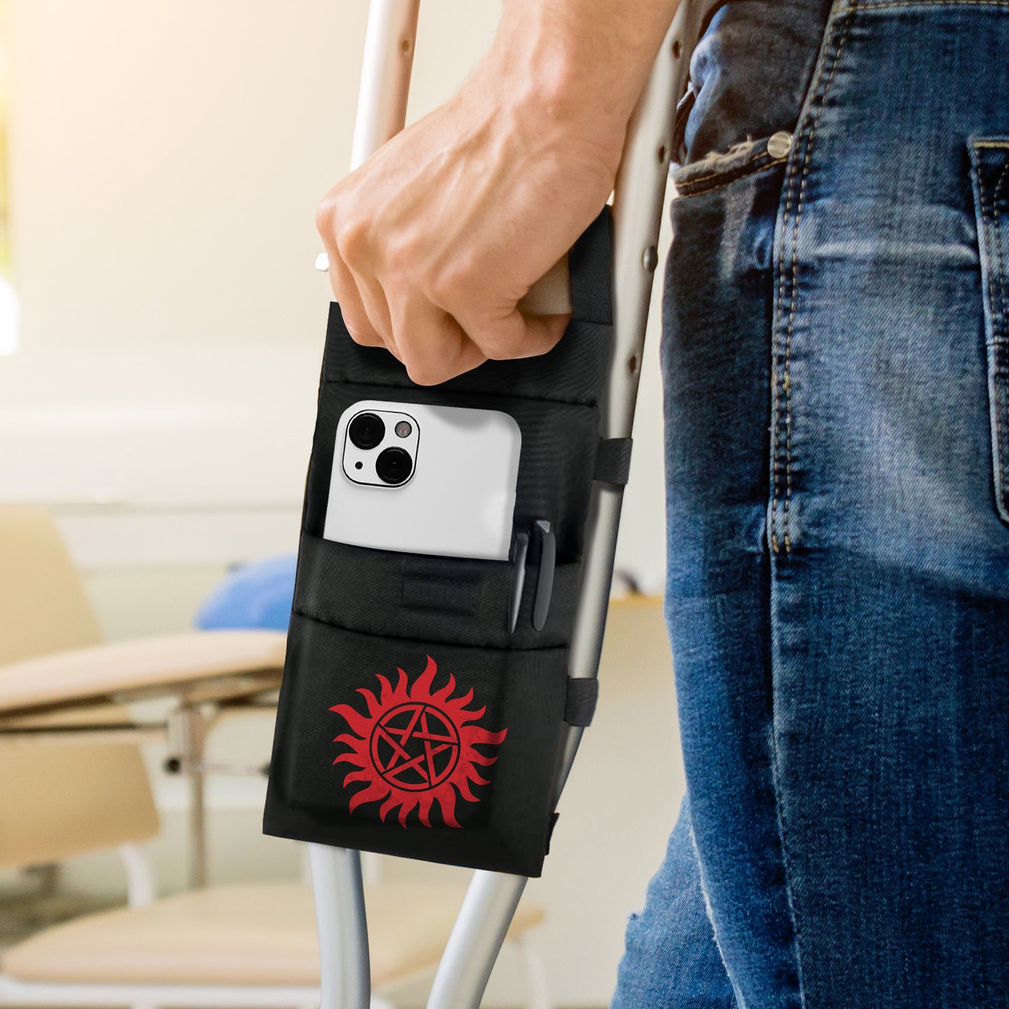 A model's leg and hand, with part of a metal crutch visible next to them. Attached to the crutch is a black nylon pouch, with the anti-possession symbol at the bottom in red. A white cell phone sits in a pocket in the pouch. In the background is a chair and desk.